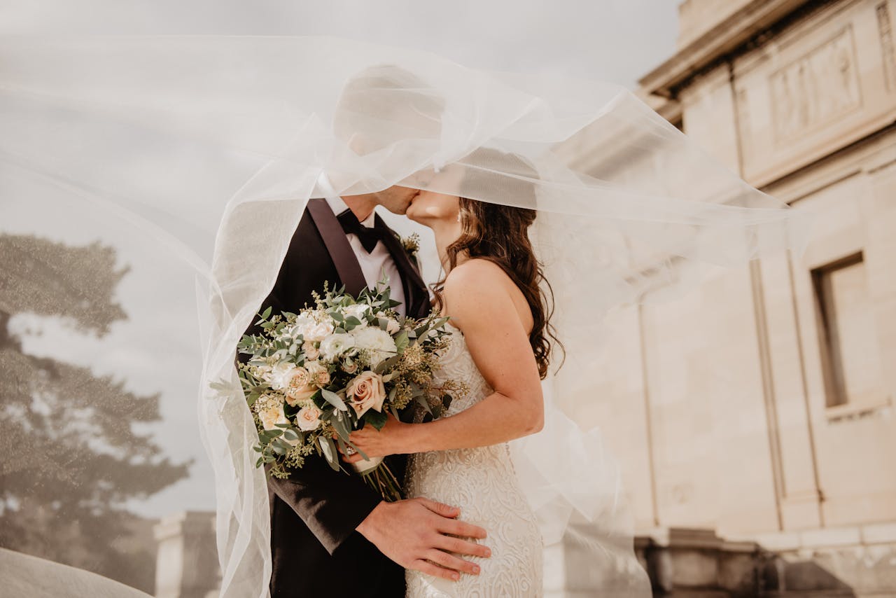 heros-img Bride and groom share a tender kiss under a veil, showcasing love and romance on their special day.